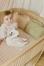 Cojín Baby Matcha junto a uno rosa en una cuna de madera, con un bebé sentado y un cojín de cisne.