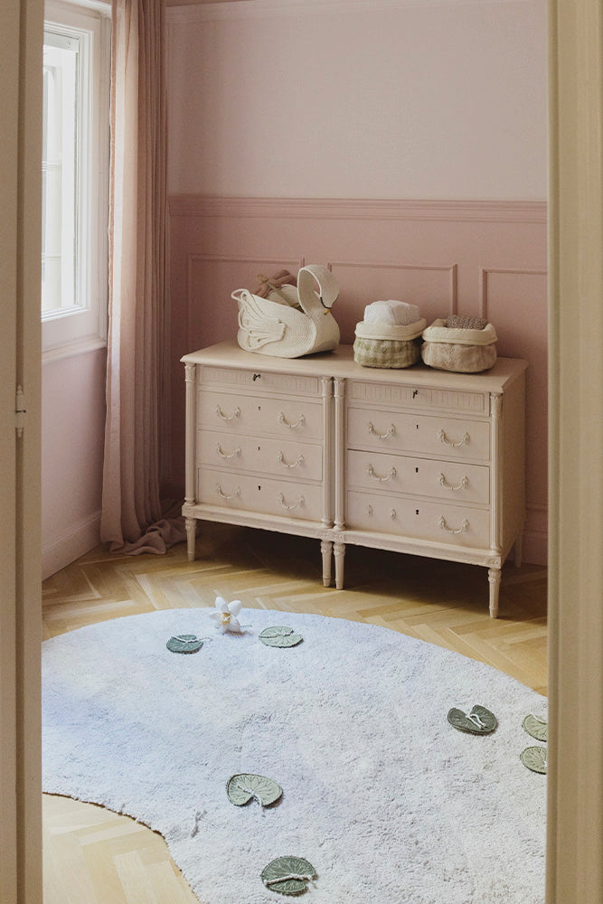 Cesta Swan sobre una cómoda en una habitación de tonos rosas, junto a cestas pequeñas y una alfombra con nenúfares.