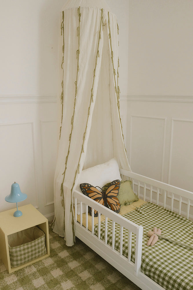 Dosel de crepé marfil sobre una cama infantil blanca con manta de cuadros.