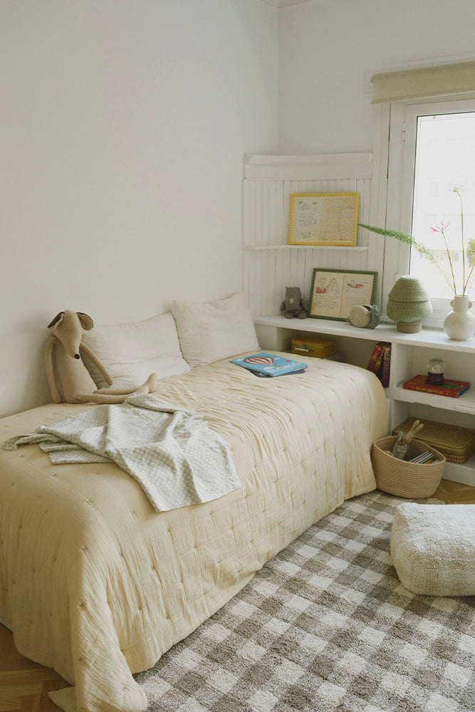 Manta Tiles azul salvia sobre una cama infantil junto a una alfombra de cuadros.