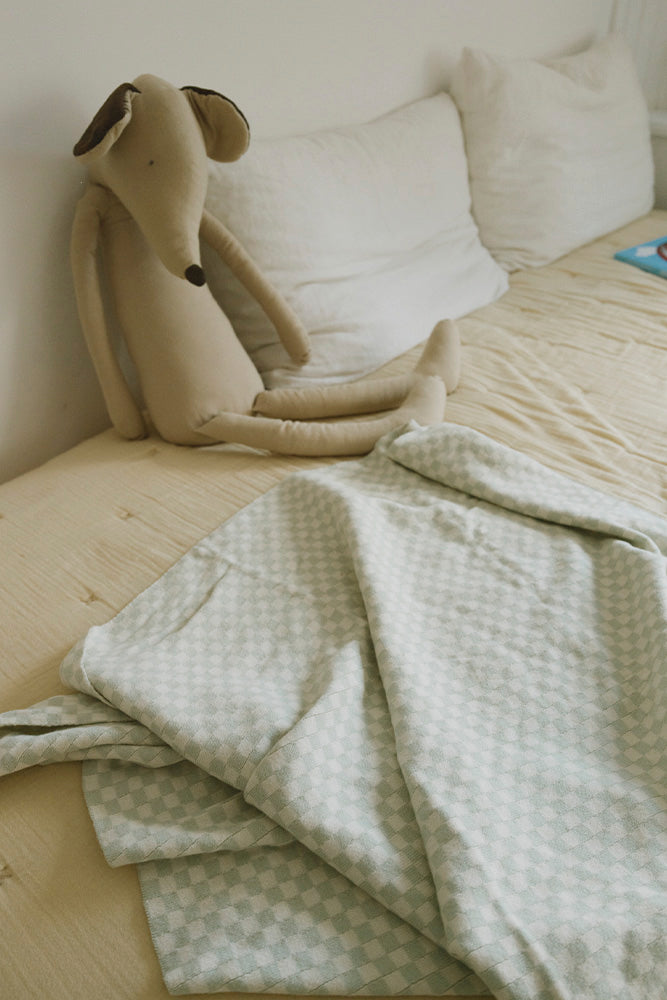 Manta Tiles azul salvia sobre una cama junto a un perro de peluche.