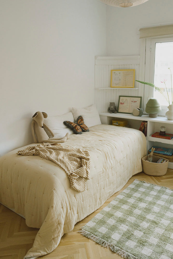 Manta Tiles miel sobre una cama infantil junto a un peluche y una alfombra de cuadros verde.