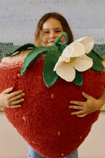 Niña abrazando el cojín Strawberry, con semillas bordadas, hojas verdes y una flor blanca.