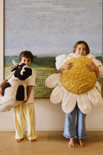Dos niños sostienen cojines decorativos: White Daisy con forma de margarita y otro con forma de vaca.