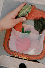Detalle de una mano colocando una verdura de tela en el bolsillo transparente con forma de olla.