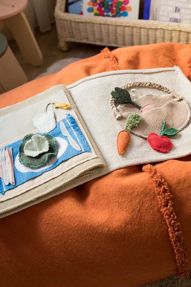 Libro textil abierto sobre una manta naranja, con un plato, cubiertos y verduras junto a una cesta.