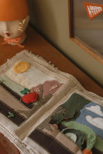 Libro sensorial abierto sobre una mesa, con páginas de tela que representan el huerto y sus verduras.